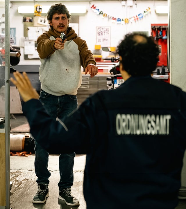 "SOKO Köln - Engel": Maximilian Angerer (Ludwig Brix) steht in einer Werkstatt und richtet mit wütendem Blick eine Waffe auf Sinan Uzun (Ali Berber), der mit dem Rücken zur Kamera vor ihm steht. Sinan hat die Arme in kapitulierender Geste nach oben gehoben. Auf seiner Jacke ist verschwommen die Aufschrift "Ordnungsamt" zu lesen. Im Hintergrund sieht man Josephine Feiler (Hanni Lorenz) auf dem Boden der Werkstatt liegen. An der Wand ist eine "Happy Birthday"-Girlande befestigt.