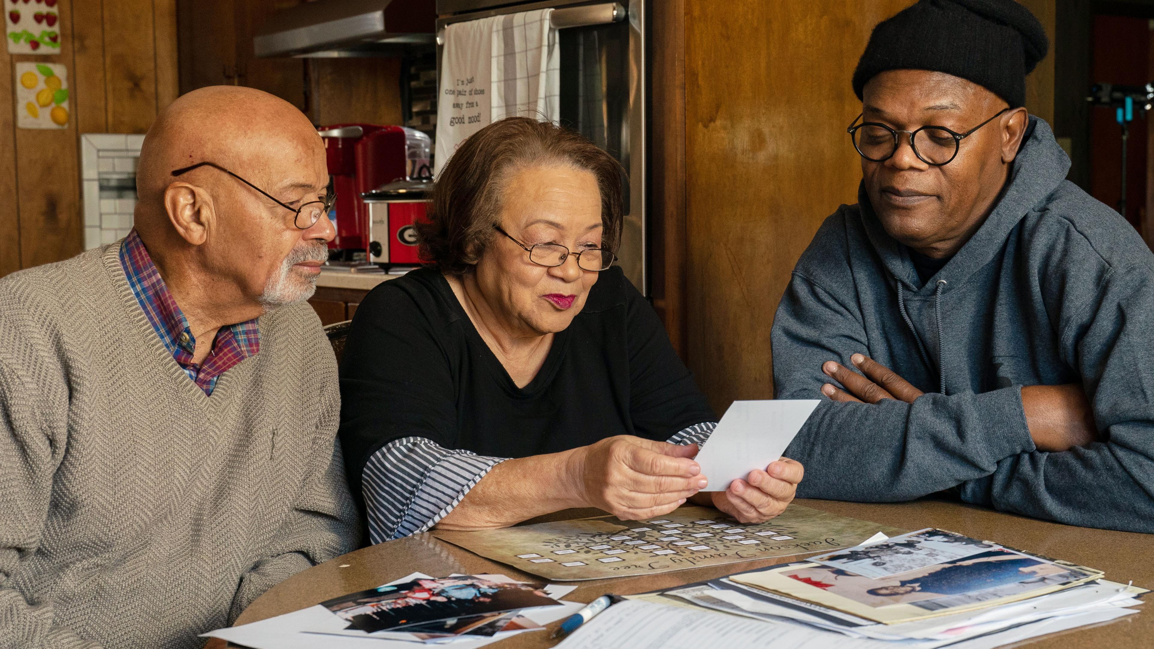 Schauspieler Samuel L. Jackson sitzt gemeinsam mit seiner Cousine Hilda und seinem Cousin Arthur an einem Tisch, auf dem Fotos und Unterlagen liegen. Hilda hält ein Foto in den Händen, auf das alle drei schauen.