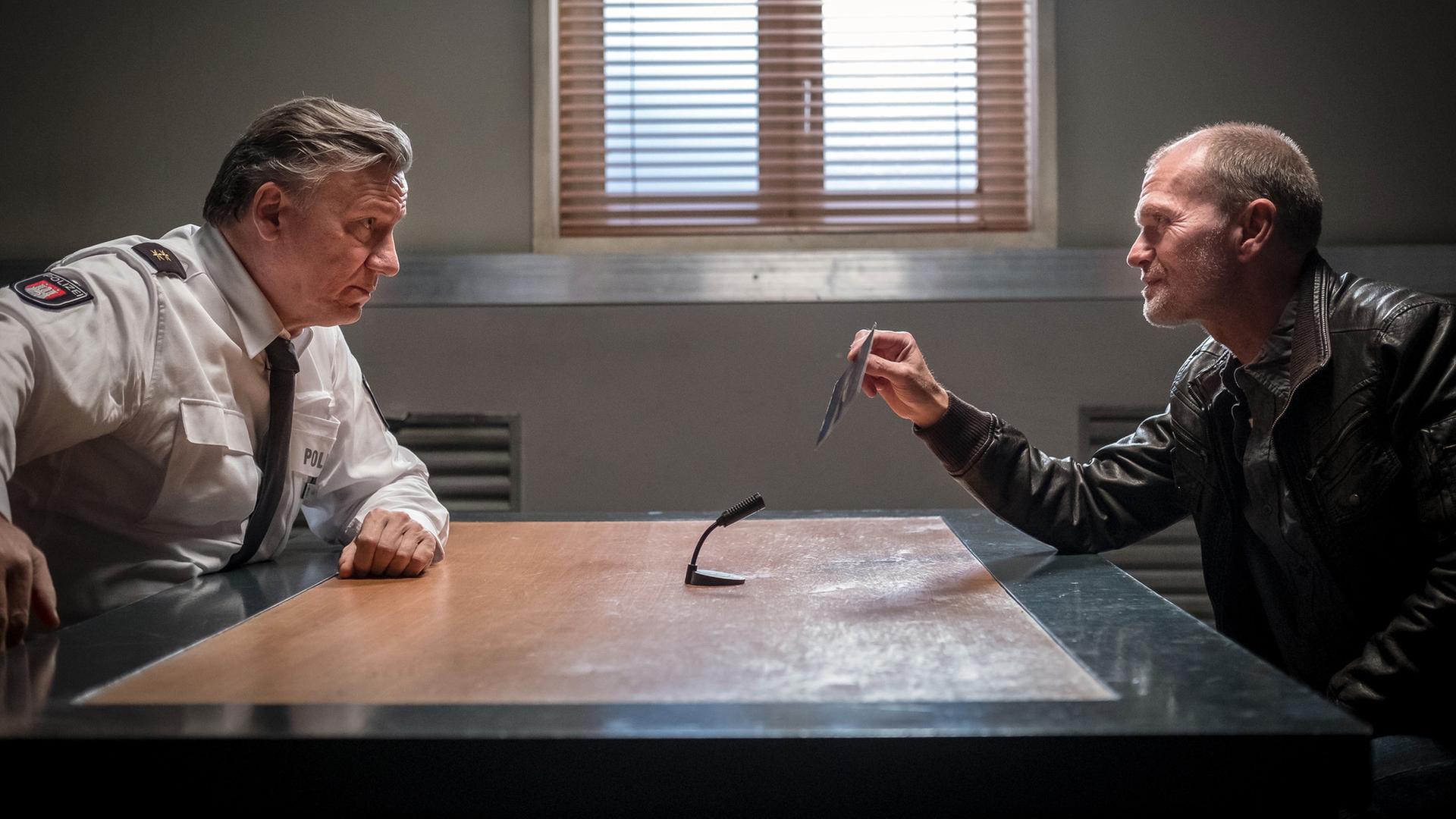 "Notruf Hafenkante - Entführt": Im Befragungsraum sitzen sich Wolf Haller (Hannes Hellmann) und Gregor Ludwig (Michael Witte) an einem Tisch gegenüber und sehen sich an. Ludwig hält ein Foto in seiner rechten Hand, das er Haller zeigt. In der Mitte des Tisches steht ein Mikrofon, das auf Ludwig gerichtet ist.