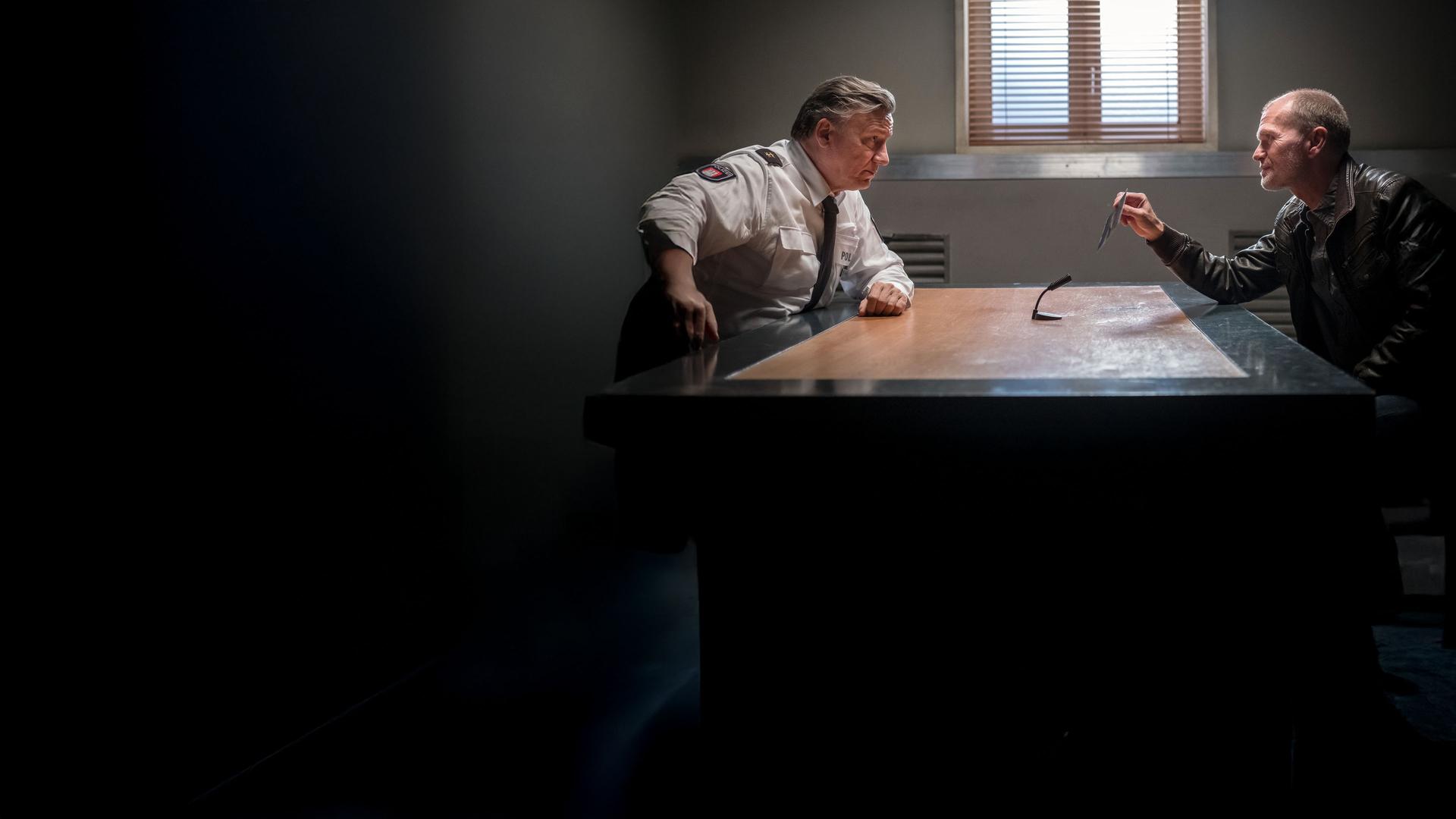 "Notruf Hafenkante - Entführt": Im Befragungsraum sitzen sich Wolf Haller (Hannes Hellmann) und Gregor Ludwig (Michael Witte) an einem Tisch gegenüber und sehen sich an. Ludwig hält ein Foto in seiner rechten Hand, das er Haller zeigt. In der Mitte des Tisches steht ein Mikrofon, das auf Ludwig gerichtet ist.