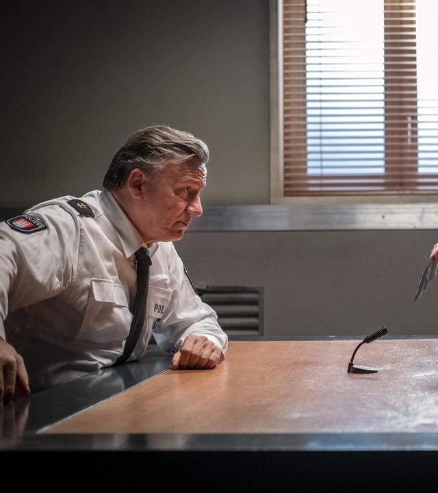 "Notruf Hafenkante - Entführt": Im Befragungsraum sitzen sich Wolf Haller (Hannes Hellmann) und Gregor Ludwig (Michael Witte) an einem Tisch gegenüber und sehen sich an. Ludwig hält ein Foto in seiner rechten Hand, das er Haller zeigt. In der Mitte des Tisches steht ein Mikrofon, das auf Ludwig gerichtet ist.