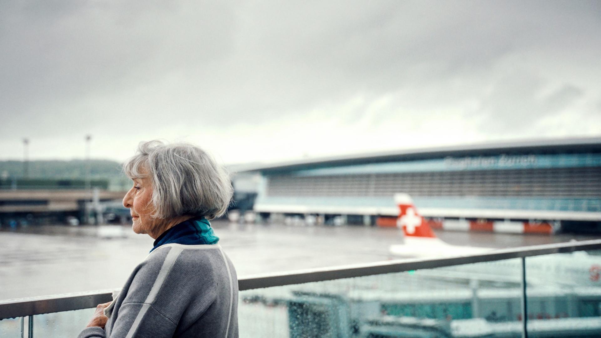 Eine Frau steht am Flughafen Zürich und blickt in die Ferne. Im Hintergrund ist ein Teil des Flughafengebäudes und ein Flugzeug mit Schweizer Flagge zu sehen.