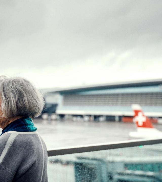 Eine Frau steht am Flughafen Zürich und blickt in die Ferne. Im Hintergrund ist ein Teil des Flughafengebäudes und ein Flugzeug mit Schweizer Flagge zu sehen.