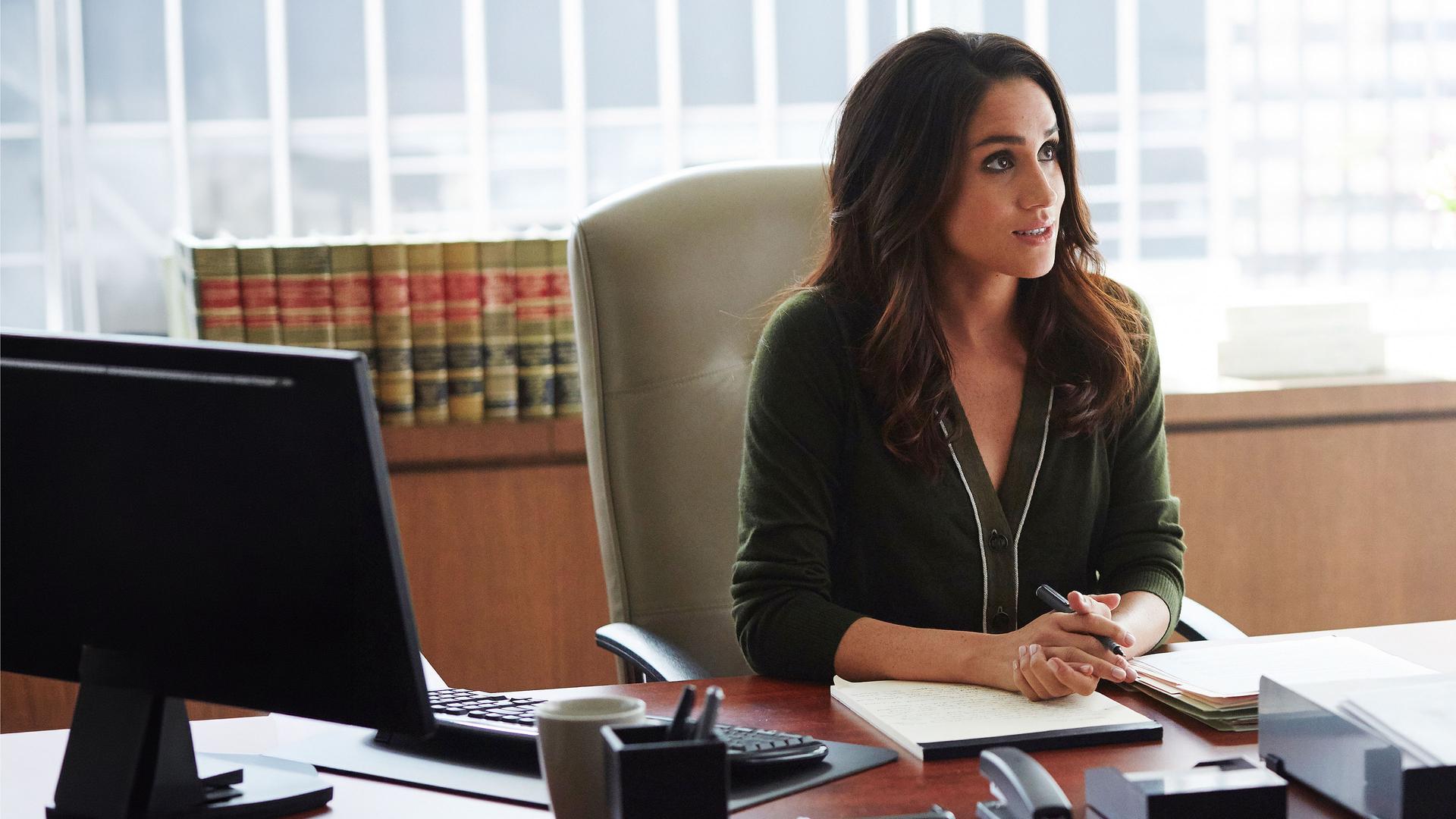 "Suits - Entgleist": Rachel (Meghan Markle) sitzt im Büro an einem Schreibtisch. Ihre Hände sind ineinandergelegt, halten einen Stift und liegen auf der Tischplatte auf einem Block.