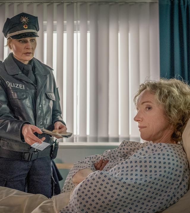 "Notruf Hafenkante - Entgleisung": Annette Drewitz (Claudia Geisler-Bading) liegt in ihrem Bett im Krankenzimmer des Elbkrankenhauses. Sie hat die Arme vor dem Oberkörper verschränkt und schaut demonstrativ zur Seite. Neben ihrem Bett steht Melanie Hansen (Sanna Englund) und hält ihr eine Visitenkarte hin.
