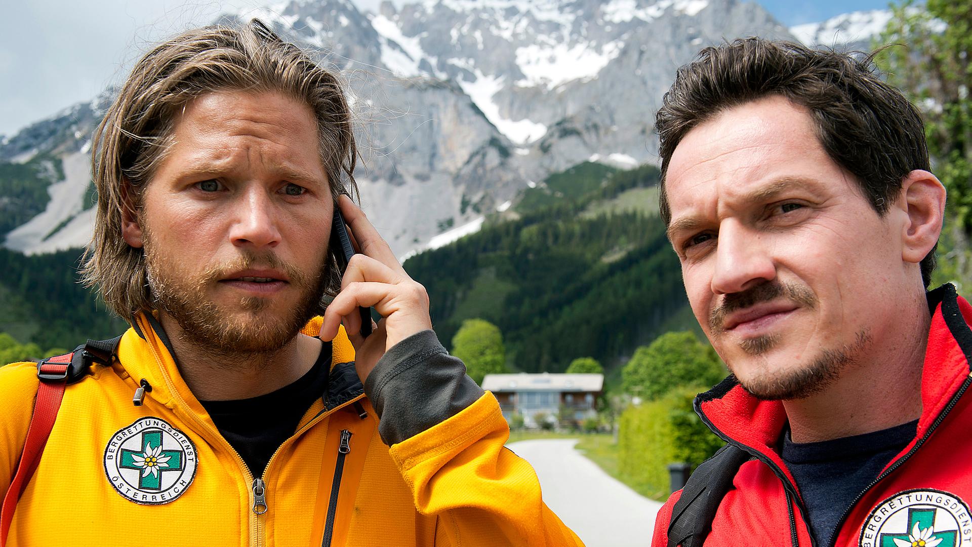  Markus (Sebastian Ströbel) steht mit dem Handy am Ohr auf der Straße. Neben ihm steht Tobias (Markus Brandl). Beide tragen ihre Bergretterjacke und blicken nachdenklich.