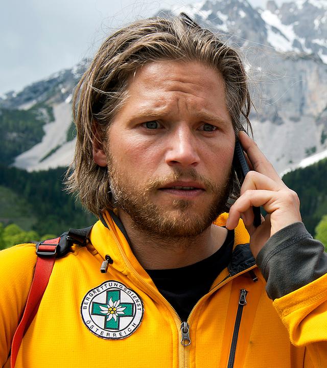  Markus (Sebastian Ströbel) steht mit dem Handy am Ohr auf der Straße. Neben ihm steht Tobias (Markus Brandl). Beide tragen ihre Bergretterjacke und blicken nachdenklich.