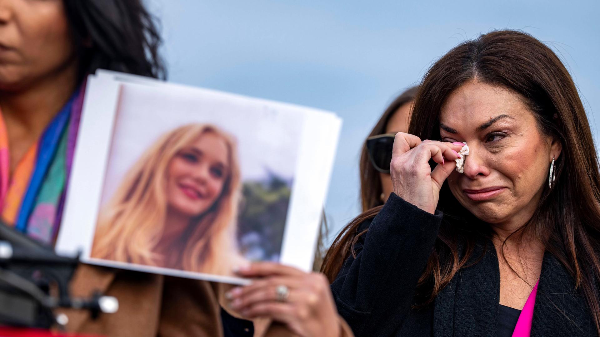 Eine Frau mit langen dunklen Haaren wischt sich mit einem Taschentuch die Tränen aus den Augen, während eine zweite das Foto einer jungen blonden Frau hoch hält.