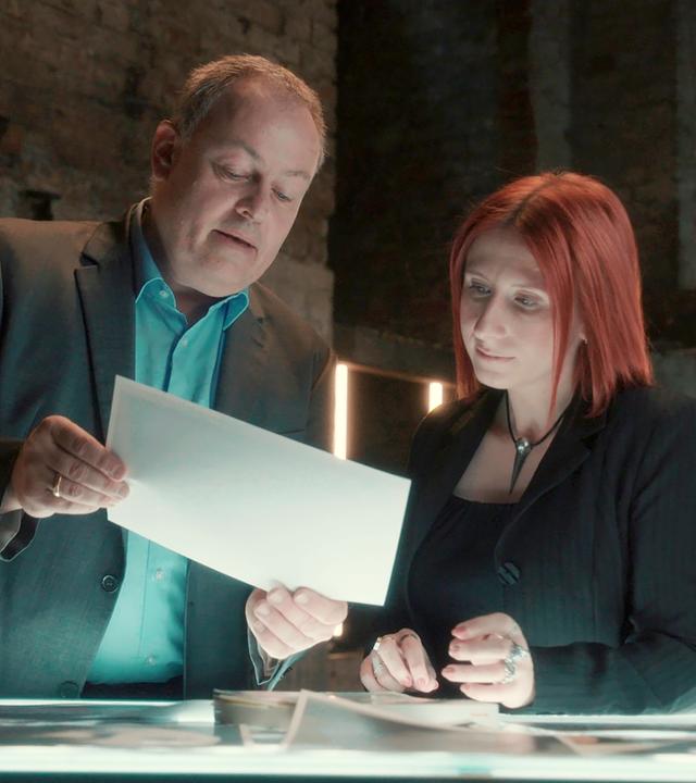 Stephan Harbort und Lydia Benecke stehen nebeneinander an einem Tisch und schauen auf ein Papier, das Harbort in seinen Händen hält.