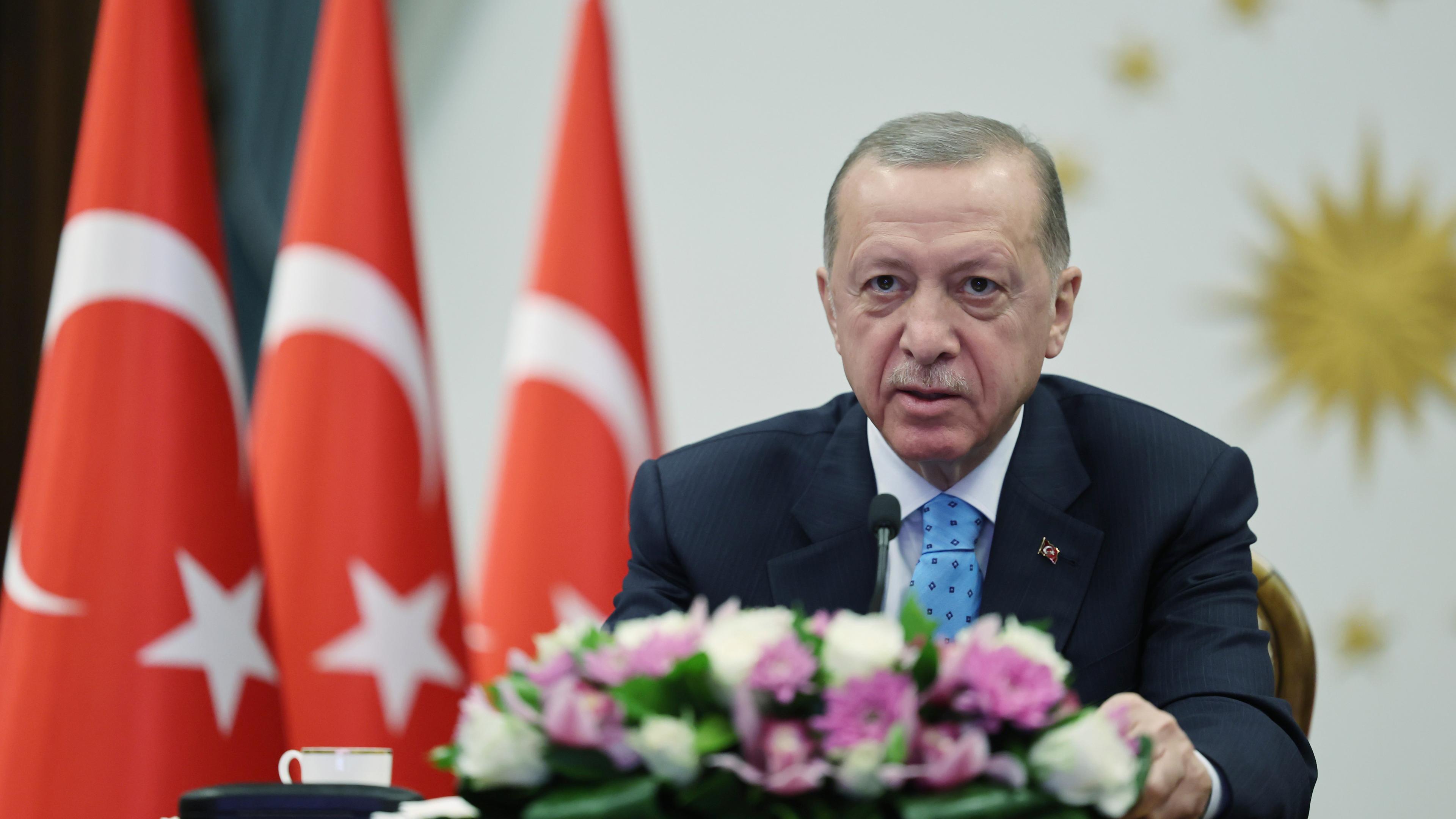 Erdogan sitzt an einem Tisch hinter einem Strauß Blumen; im Hintergrund Türkei-Flaggen.