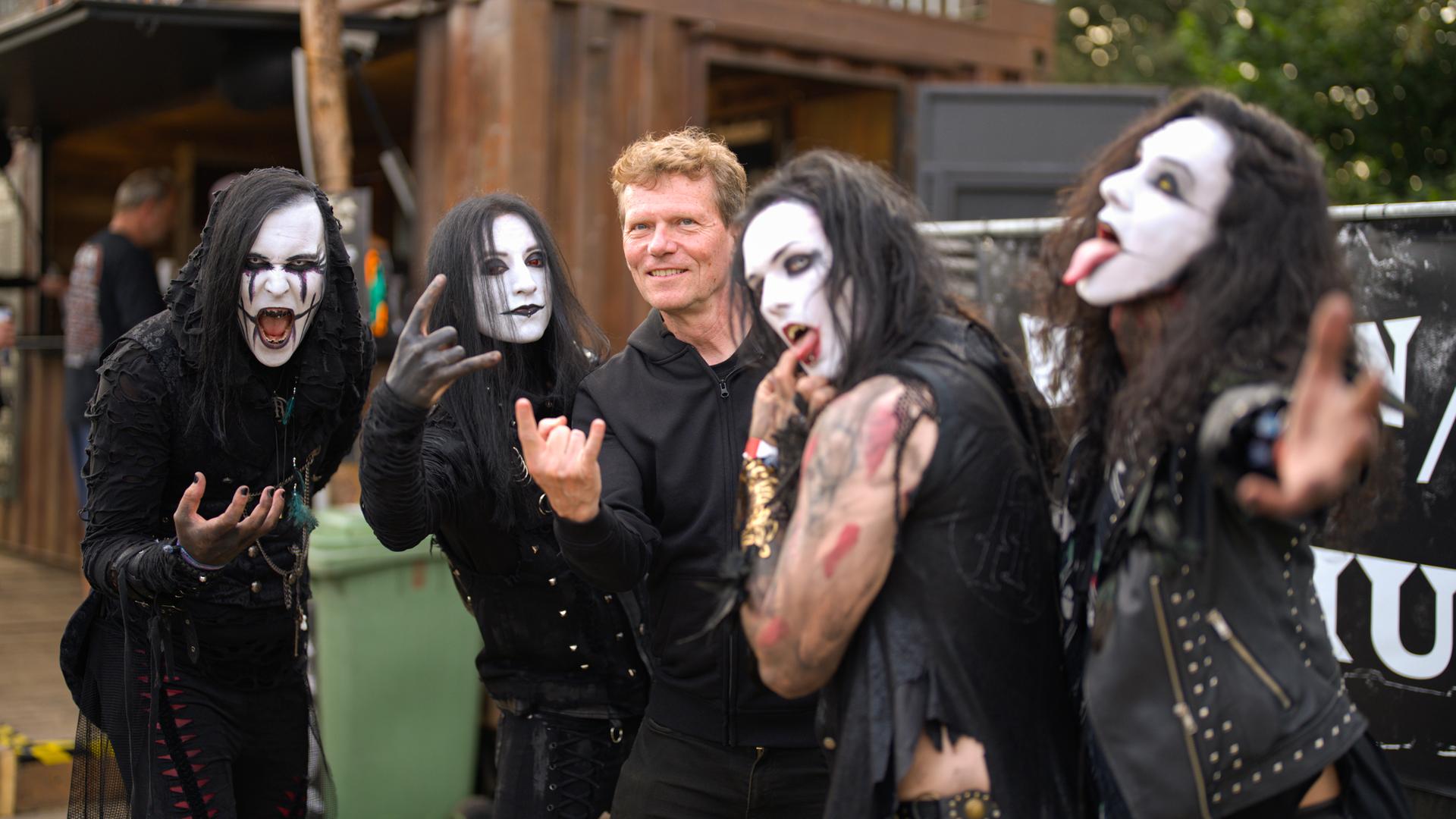 Ein Mann mit heller Haut und kurzen rötlichen Haaren steht zwischen vier weiß geschminkten Gestalten mit schwarzen langen Haaren. Alle machen typische Metal-Gesten.