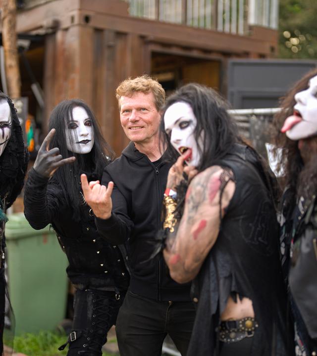 Ein Mann mit heller Haut und kurzen rötlichen Haaren steht zwischen vier weiß geschminkten Gestalten mit schwarzen langen Haaren. Alle machen typische Metal-Gesten.