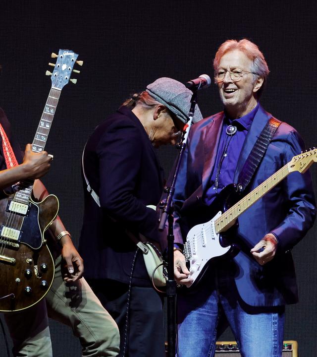 Eric Clapton steht mit seiner Gitarre und einem weiteren Musiker mit Gitarre auf der Bühne.