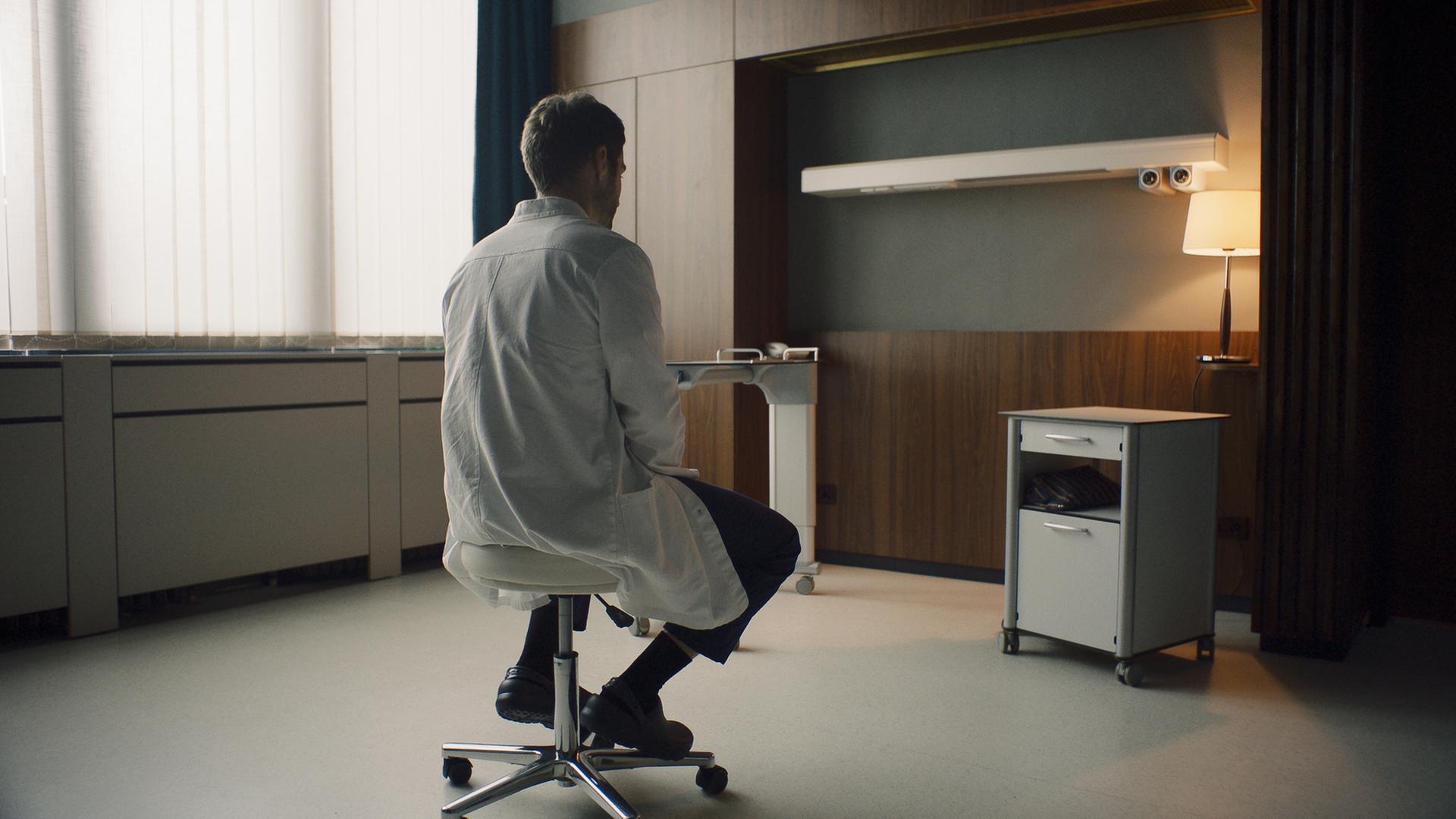"Doktor Ballouz - Erinnerungen": Dr. Mark Schilling (Daniel Fritz) sitzt in einem Patientenzimmer, das nach dem Tod des Patienten leergeräumt wurde, nachdenklich auf einem Hocker.