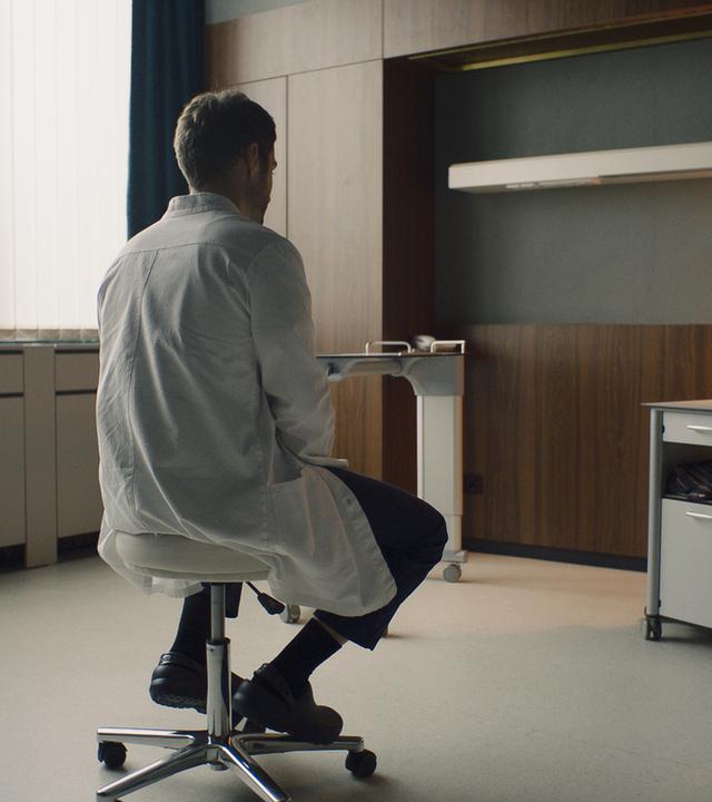 "Doktor Ballouz - Erinnerungen": Dr. Mark Schilling (Daniel Fritz) sitzt in einem Patientenzimmer, das nach dem Tod des Patienten leergeräumt wurde, nachdenklich auf einem Hocker.