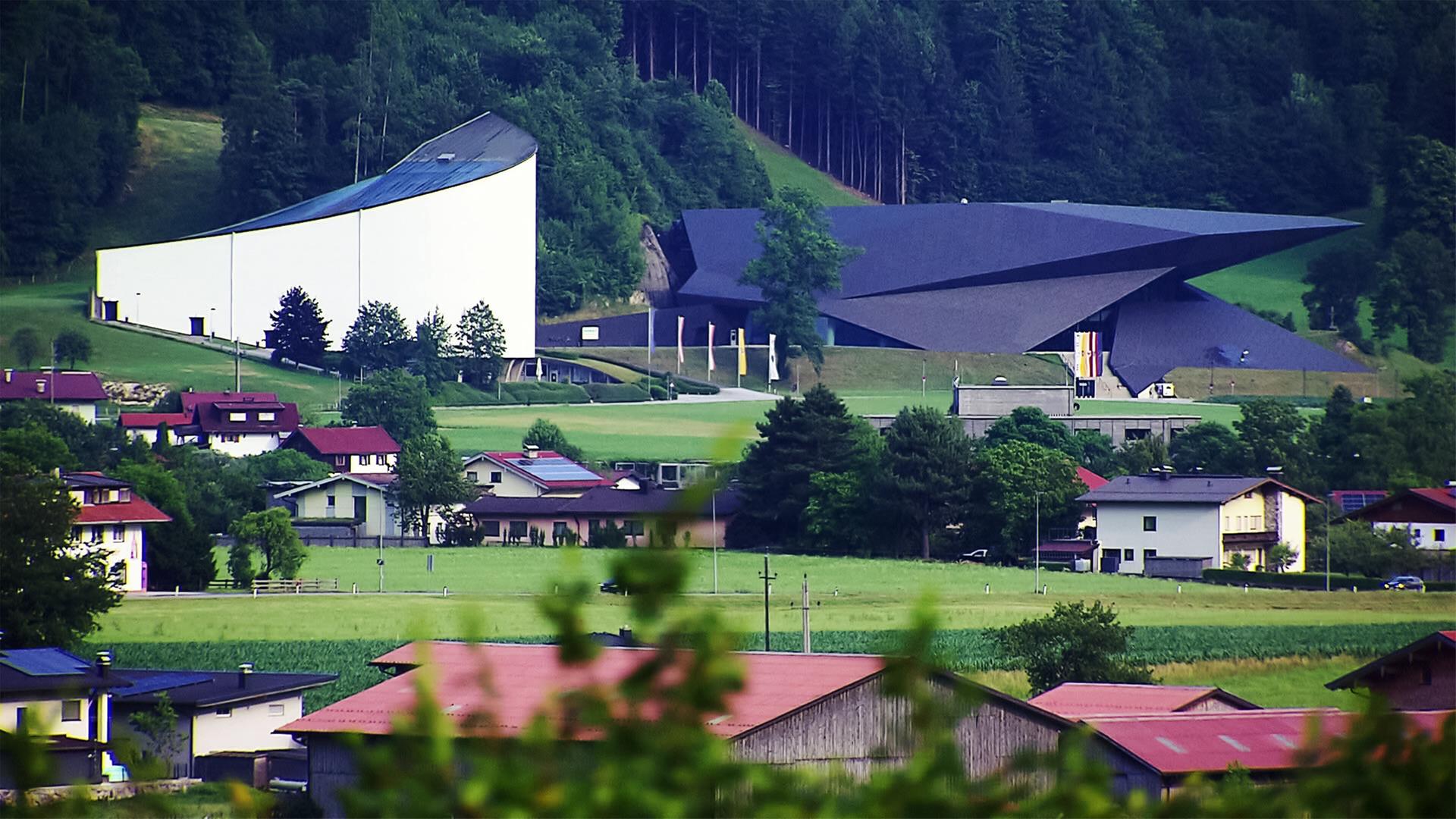 Das Bild zeigt das Festspielhaus Erl, ein markantes architektonisches Gebäude, das in eine grüne Landschaft integriert ist. Es befindet sich im Tiroler Dorf Erl, nahe der bayerischen Grenze. Auf der linken Seite sieht man die hellere, geschwungene Fassade des Festspielhauses, die sich harmonisch in die Umgebung einfügt. Daneben befindet sich ein dunkleres Gebäude mit einer ausgefallenen, spitzen Dachform.   Im Vordergrund sind mehrere Wohnhäuser mit roten Dächern und Gärten zu erkennen, die in einer hügeligen Landschaft angeordnet sind. Die Umgebung ist geprägt von grünen Wiesen, Bäumen und sanften Hügeln, die die Idylle des ländlichen Bereichs unterstreichen. Im Hintergrund sind dichte Wälder zu sehen, die sich bis zu den Gipfeln der Berge hochziehen.   Das Bild vermittelt einen Eindruck von der ruhigen Atmosphäre und der kulturellen Bedeutung der Tiroler Festspiele, die in dieser malerischen Umgebung stattfinden.