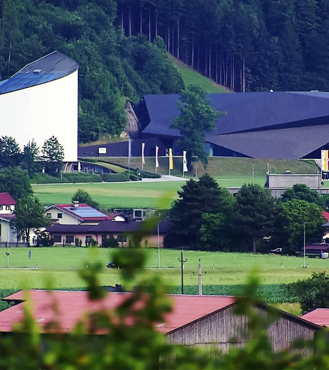 Das Bild zeigt das Festspielhaus Erl, ein markantes architektonisches Gebäude, das in eine grüne Landschaft integriert ist. Es befindet sich im Tiroler Dorf Erl, nahe der bayerischen Grenze. Auf der linken Seite sieht man die hellere, geschwungene Fassade des Festspielhauses, die sich harmonisch in die Umgebung einfügt. Daneben befindet sich ein dunkleres Gebäude mit einer ausgefallenen, spitzen Dachform.   Im Vordergrund sind mehrere Wohnhäuser mit roten Dächern und Gärten zu erkennen, die in einer hügeligen Landschaft angeordnet sind. Die Umgebung ist geprägt von grünen Wiesen, Bäumen und sanften Hügeln, die die Idylle des ländlichen Bereichs unterstreichen. Im Hintergrund sind dichte Wälder zu sehen, die sich bis zu den Gipfeln der Berge hochziehen.   Das Bild vermittelt einen Eindruck von der ruhigen Atmosphäre und der kulturellen Bedeutung der Tiroler Festspiele, die in dieser malerischen Umgebung stattfinden.