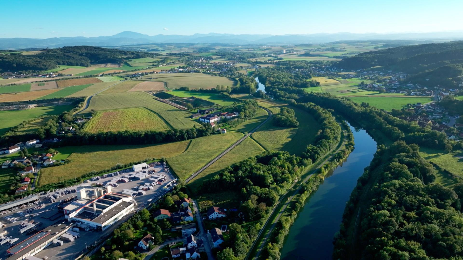 Das Bild zeigt eine weitläufige Landschaft im Erlauftal, das von sanften Hügeln und Feldern geprägt ist. Im Vordergrund sind Wege und eine kleine Siedlung zu sehen, die aus mehreren Wohnhäusern besteht. Ein Fluss, vermutlich die Erlauf, schlängelt sich durch die grüne Landschaft, flankiert von Bäumen.   In der Ferne erheben sich Berge, die die Szene umrahmen und einen klaren Himmel über der Region zeigen. Auf der linken Seite des Bildes sind landwirtschaftliche Felder in verschiedenen Grüntönen zu erkennen, während auf der rechten Seite ein größerer Parkplatz und ein Industriegebäude sichtbar sind.   Zusammen vermittelt das Bild einen Eindruck von der Kombination aus Natur und menschlicher Besiedlung, die für das Mostviertel charakteristisch ist.