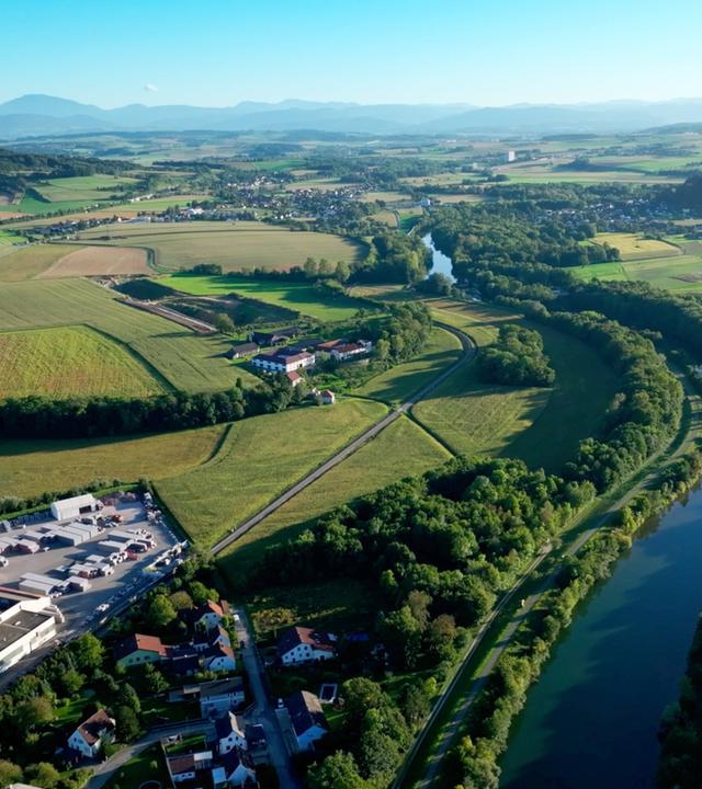 Das Bild zeigt eine weitläufige Landschaft im Erlauftal, das von sanften Hügeln und Feldern geprägt ist. Im Vordergrund sind Wege und eine kleine Siedlung zu sehen, die aus mehreren Wohnhäusern besteht. Ein Fluss, vermutlich die Erlauf, schlängelt sich durch die grüne Landschaft, flankiert von Bäumen.   In der Ferne erheben sich Berge, die die Szene umrahmen und einen klaren Himmel über der Region zeigen. Auf der linken Seite des Bildes sind landwirtschaftliche Felder in verschiedenen Grüntönen zu erkennen, während auf der rechten Seite ein größerer Parkplatz und ein Industriegebäude sichtbar sind.   Zusammen vermittelt das Bild einen Eindruck von der Kombination aus Natur und menschlicher Besiedlung, die für das Mostviertel charakteristisch ist.