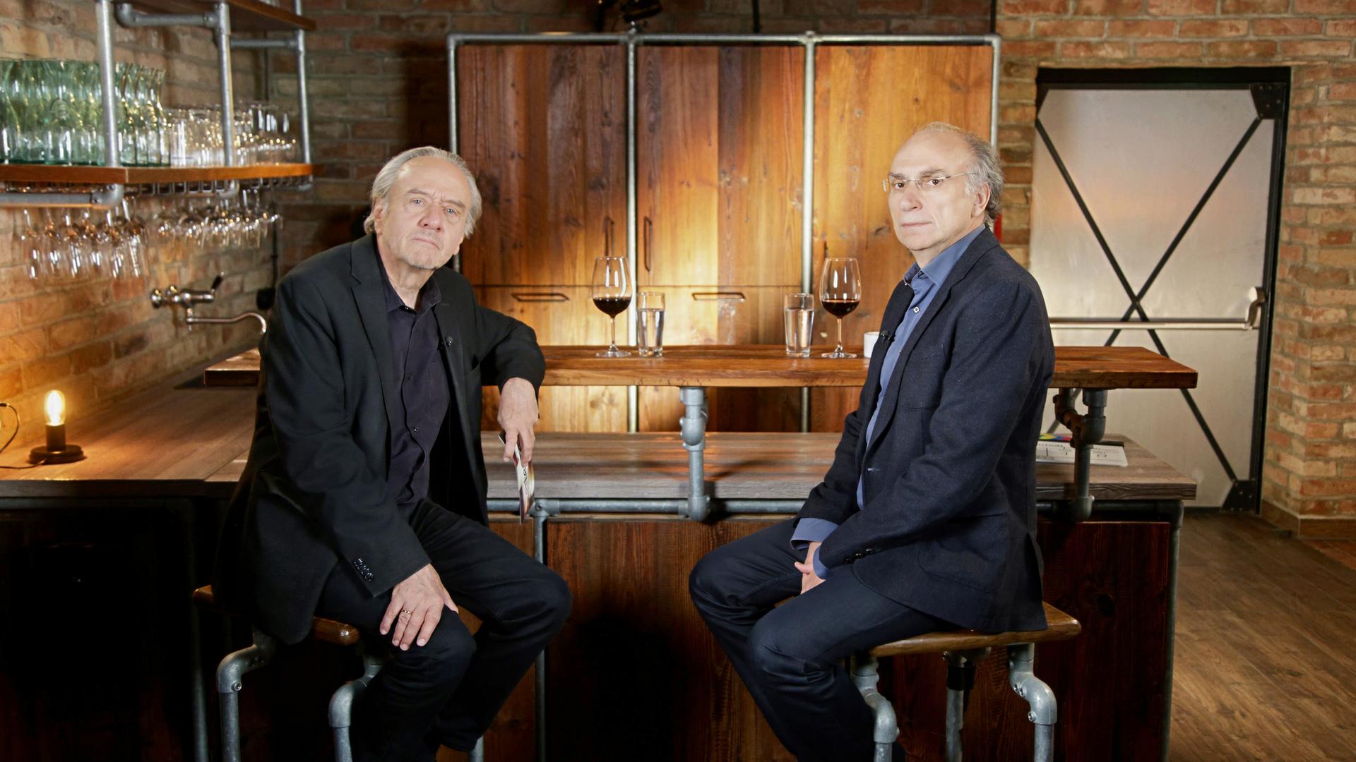 Das Bild zeigt zwei Männer, die an einer Bartheke sitzen. Der Raum hat eine rustikale Atmosphäre mit Backsteinmauern und Holzelementen. Die Bartheke ist aus Holz und Metallrohren gefertigt. Auf der Theke stehen Gläser, darunter ein Glas mit rotem Wein und ein weiteres mit Wasser.   Der Mann links trägt einen schwarzen Anzug mit einem schwarzen Hemd und hat graues Haar. Er blickt direkt in die Kamera, wirkt nachdenklich. Der Mann rechts sitzt in einem dunklen, strukturierten Blazer und einem blauen Hemd. Er hat kurze, blonde Haare und trägt eine Brille. Auch er schaut in die Kamera, hat einen ernsten Gesichtsausdruck.   Im Hintergrund sind weitere Regale mit Gläsern zu sehen, und an der Wand hängen Lampen, die für indirektes Licht sorgen. In der Ecke ist eine Tür zu erkennen, die einen schlichten Design hat. Die Atmosphäre des Raumes wirkt freundlich und einladend, geeignet für Gespräche oder Interviews.