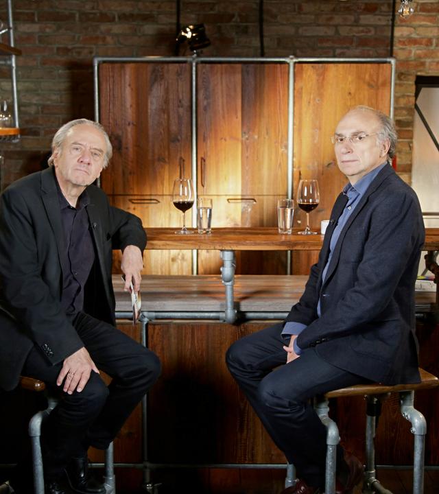 Das Bild zeigt zwei Männer, die an einer Bartheke sitzen. Der Raum hat eine rustikale Atmosphäre mit Backsteinmauern und Holzelementen. Die Bartheke ist aus Holz und Metallrohren gefertigt. Auf der Theke stehen Gläser, darunter ein Glas mit rotem Wein und ein weiteres mit Wasser.   Der Mann links trägt einen schwarzen Anzug mit einem schwarzen Hemd und hat graues Haar. Er blickt direkt in die Kamera, wirkt nachdenklich. Der Mann rechts sitzt in einem dunklen, strukturierten Blazer und einem blauen Hemd. Er hat kurze, blonde Haare und trägt eine Brille. Auch er schaut in die Kamera, hat einen ernsten Gesichtsausdruck.   Im Hintergrund sind weitere Regale mit Gläsern zu sehen, und an der Wand hängen Lampen, die für indirektes Licht sorgen. In der Ecke ist eine Tür zu erkennen, die einen schlichten Design hat. Die Atmosphäre des Raumes wirkt freundlich und einladend, geeignet für Gespräche oder Interviews.