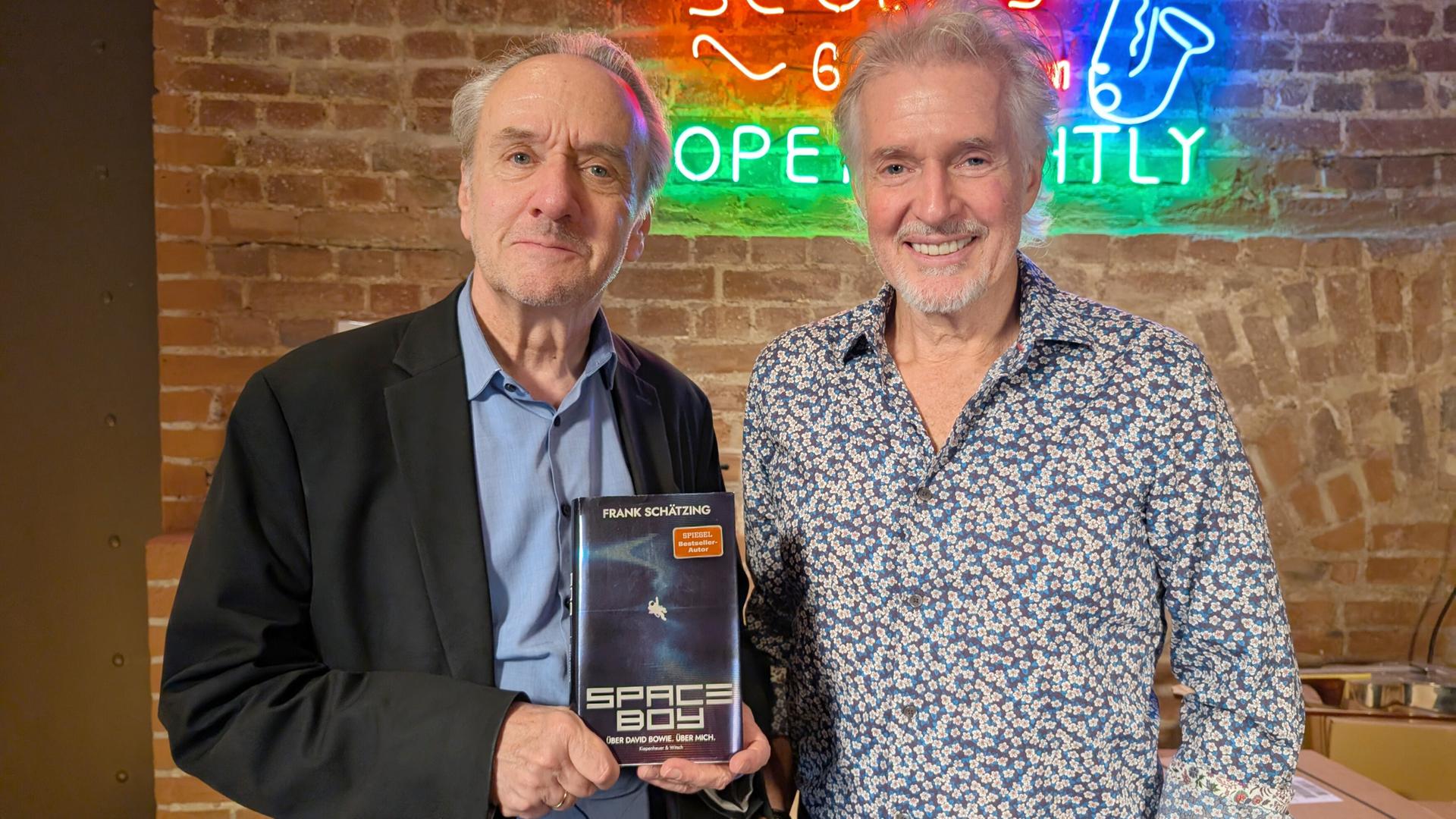 Das Bild zeigt zwei Männer, die in einem Tonstudio stehen. Im Hintergrund erkennt man eine Wand aus Ziegelsteinen, die mit einem bunten Neon-Schild dekoriert ist. Das Neonlicht zeigt den Schriftzug „ronnie scott's“, umgeben von weiteren bunten Lichtern.  Der Mann links, Frank Schätzing, hält ein Buch mit dem Titel "Space Boy", das sich auf seine Verbindung zur Musiklegende David Bowie bezieht. Er trägt ein schwarzes Jackett über einem blauen Hemd. Sein Gesicht wirkt ernst, und er schaut direkt in die Kamera.  Der Mann rechts, Heinz Sichrovsky, lächelt und trägt ein gemustertes Hemd in Blau mit kleinen weißen Blumen. Beide Männer stehen nahe beieinander und posieren für das Foto. Im Vordergrund sind offenbar verschiedene Kartons zu sehen, was auf eine Veranstaltung oder ein Interview hinweisen könnte.   Die Atmosphäre des Raumes wirkt kreativ und einladend, passend zu einem Tonstudio.