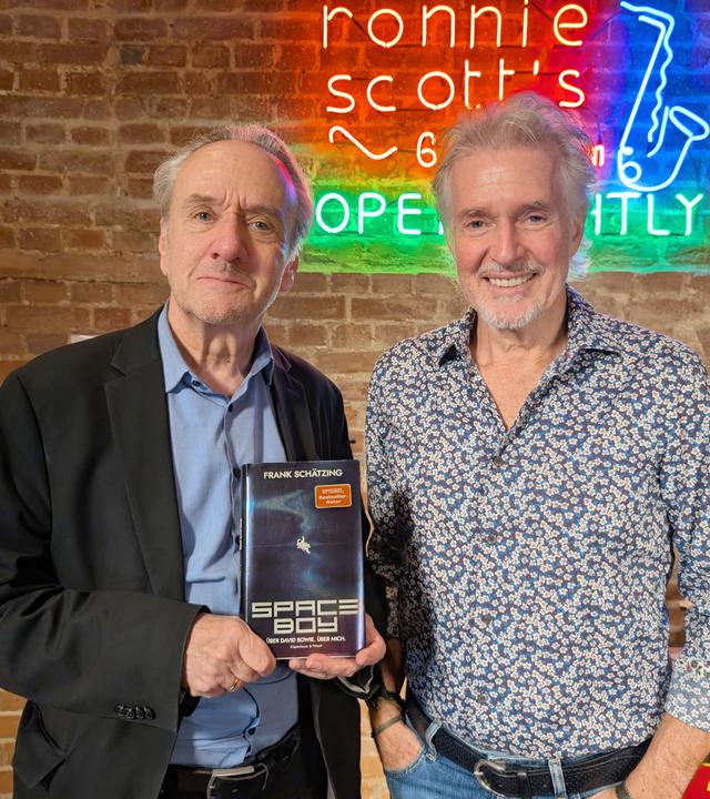 Das Bild zeigt zwei Männer, die in einem Tonstudio stehen. Im Hintergrund erkennt man eine Wand aus Ziegelsteinen, die mit einem bunten Neon-Schild dekoriert ist. Das Neonlicht zeigt den Schriftzug „ronnie scott's“, umgeben von weiteren bunten Lichtern.  Der Mann links, Frank Schätzing, hält ein Buch mit dem Titel "Space Boy", das sich auf seine Verbindung zur Musiklegende David Bowie bezieht. Er trägt ein schwarzes Jackett über einem blauen Hemd. Sein Gesicht wirkt ernst, und er schaut direkt in die Kamera.  Der Mann rechts, Heinz Sichrovsky, lächelt und trägt ein gemustertes Hemd in Blau mit kleinen weißen Blumen. Beide Männer stehen nahe beieinander und posieren für das Foto. Im Vordergrund sind offenbar verschiedene Kartons zu sehen, was auf eine Veranstaltung oder ein Interview hinweisen könnte.   Die Atmosphäre des Raumes wirkt kreativ und einladend, passend zu einem Tonstudio.