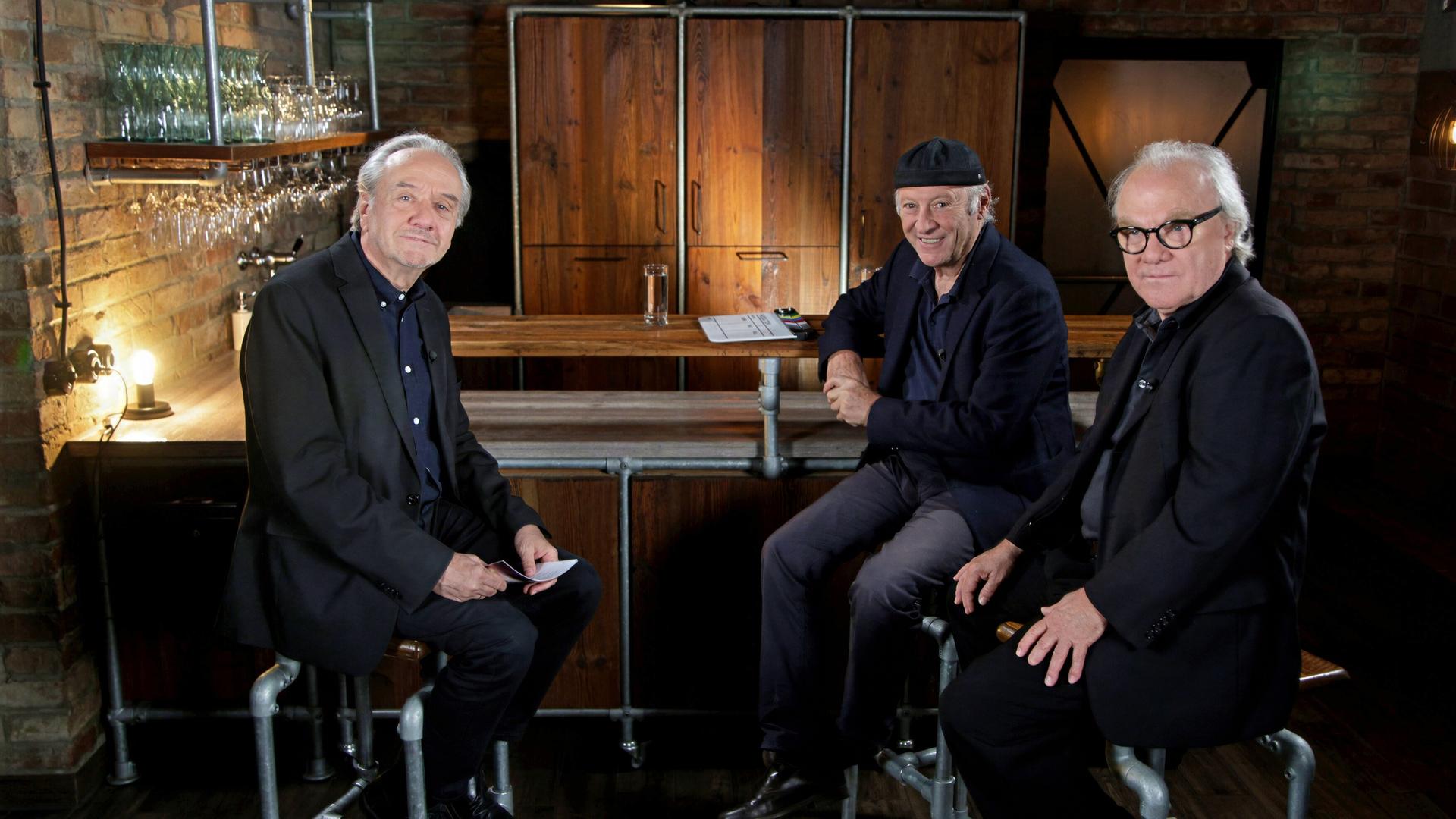 Das Bild zeigt drei Männer, die in einem gemütlich gestalteten Innenraum sitzen. Der Hintergrund besteht aus einer Steinwand, die mit Holzregalen dekoriert ist, auf denen Gläser stehen. In der Mitte des Bildes verläuft eine Bar aus Holz, hinter der sich eine geschlossene Holzschranktür befindet.   Die drei Männer sind auf Barhockern platziert. Der Mann links hat graue Haare, trägt einen dunklen Anzug und schaut freundlich in die Kamera. Der Mann in der Mitte trägt ein schwarzes Hemd und eine schwarze Kappe, lächelt und hat seine Hände auf der Tischkante abgelegt. Der Mann rechts trägt eine dunkle Brille und eine schwarze Jacke. Er sitzt mit verschränkten Armen, hat einen ernsten, aber freundlichen Gesichtsausdruck.  Die Beleuchtung ist warm und sorgt für eine einladende Atmosphäre. Insgesamt vermittelt das Bild den Eindruck eines informellen Gesprächs zwischen den drei Männern.