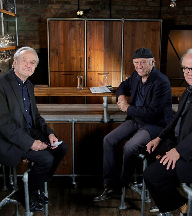 Das Bild zeigt drei Männer, die in einem gemütlich gestalteten Innenraum sitzen. Der Hintergrund besteht aus einer Steinwand, die mit Holzregalen dekoriert ist, auf denen Gläser stehen. In der Mitte des Bildes verläuft eine Bar aus Holz, hinter der sich eine geschlossene Holzschranktür befindet.   Die drei Männer sind auf Barhockern platziert. Der Mann links hat graue Haare, trägt einen dunklen Anzug und schaut freundlich in die Kamera. Der Mann in der Mitte trägt ein schwarzes Hemd und eine schwarze Kappe, lächelt und hat seine Hände auf der Tischkante abgelegt. Der Mann rechts trägt eine dunkle Brille und eine schwarze Jacke. Er sitzt mit verschränkten Armen, hat einen ernsten, aber freundlichen Gesichtsausdruck.  Die Beleuchtung ist warm und sorgt für eine einladende Atmosphäre. Insgesamt vermittelt das Bild den Eindruck eines informellen Gesprächs zwischen den drei Männern.