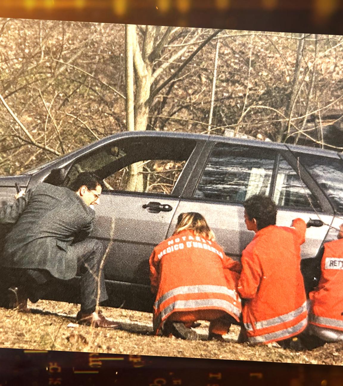 Ermittler Alexander Zelger, drei Rettungskräfte in orangefarbenen Anzügen und ein weiterer Mann gehen hinter einem Auto, das im Wald steht, in Deckung.