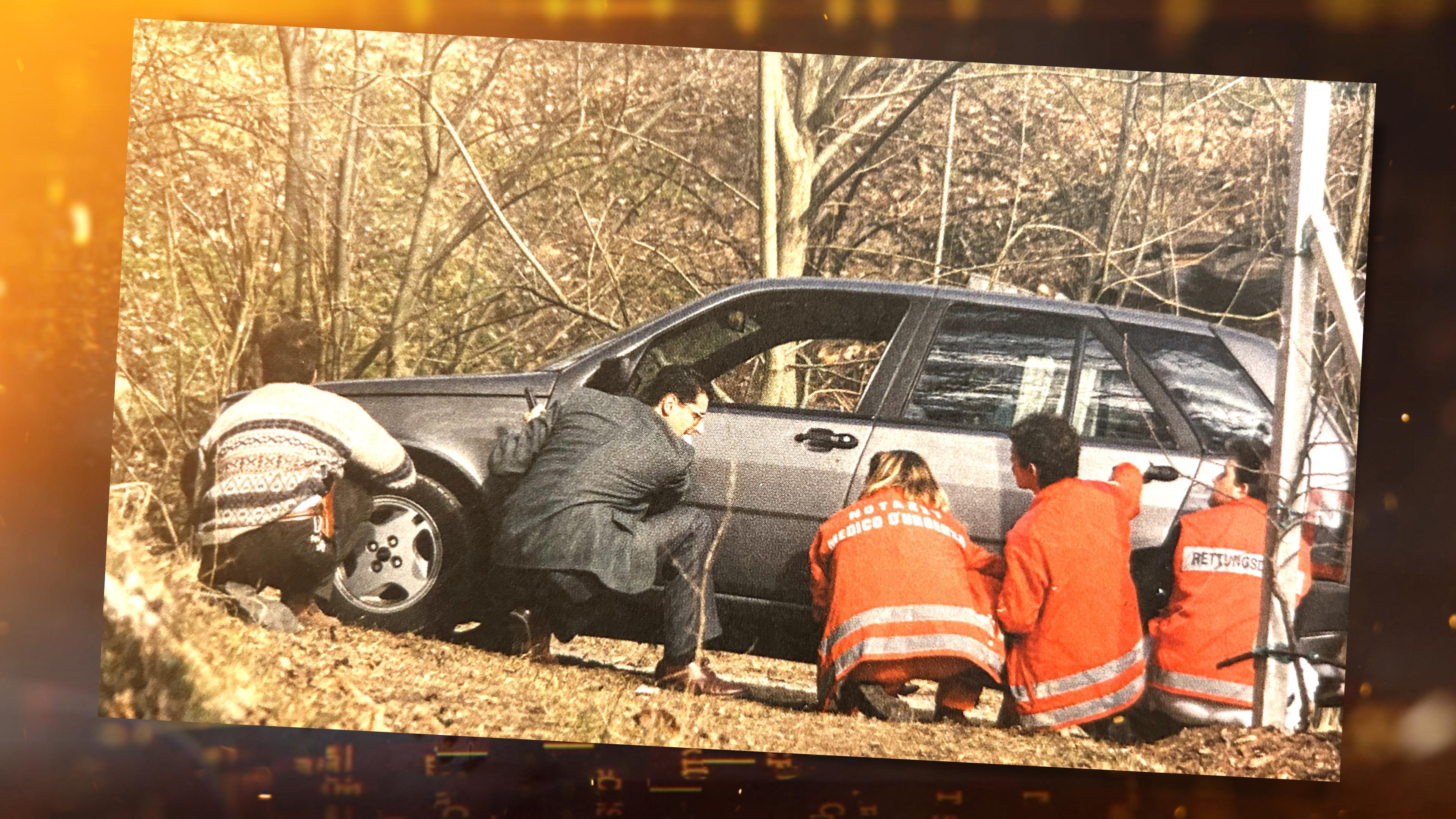 Ermittler Alexander Zelger, drei Rettungskräfte in orangefarbenen Anzügen und ein weiterer Mann gehen hinter einem Auto, das im Wald steht, in Deckung.