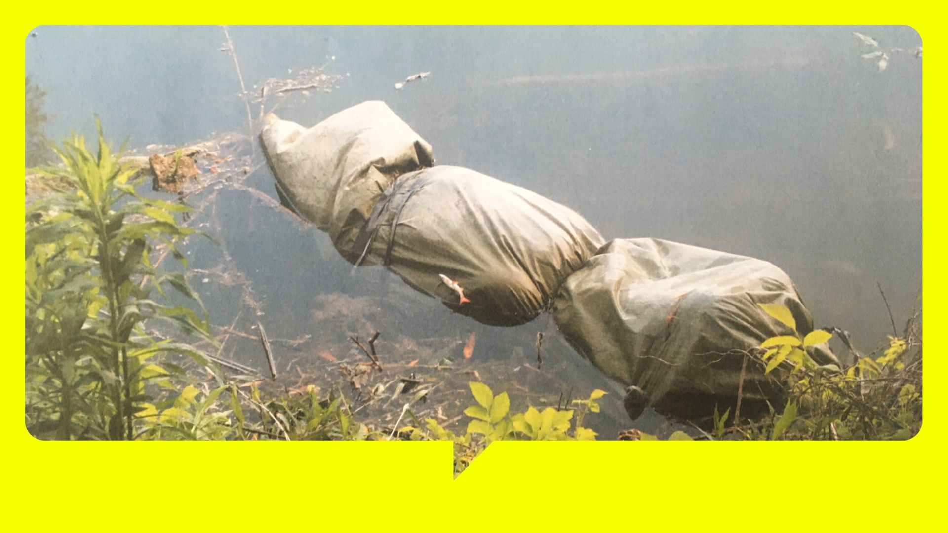 Ein mit mehreren Seilen verschnürtes Bündel treibt am Ufer eines Sees im Wasser. Form und Größe entsprechen etwa einem menschlichen Körper. Pflanzenbewuchs am Ufer. Schemenhaft ist der Grund des Sees zu erkennen.