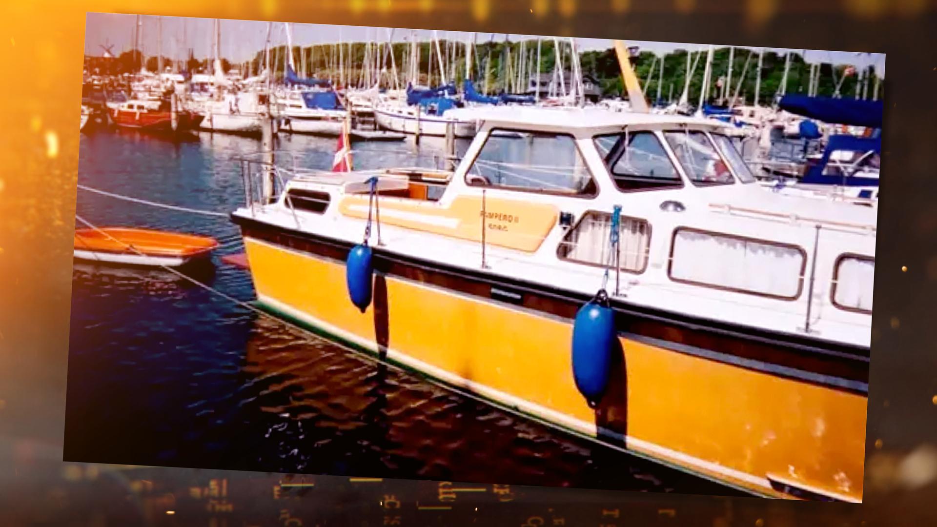 Ein weiß-gelbes Motorboot mit Kajüte liegt im Hafen. Das Boot trägt die dänische Flagge, im Hintergrund liegen weitere Segelboote.