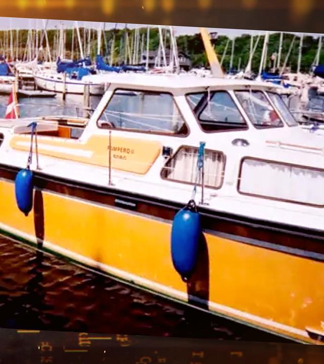 Ein weiß-gelbes Motorboot mit Kajüte liegt im Hafen. Das Boot trägt die dänische Flagge, im Hintergrund liegen weitere Segelboote.