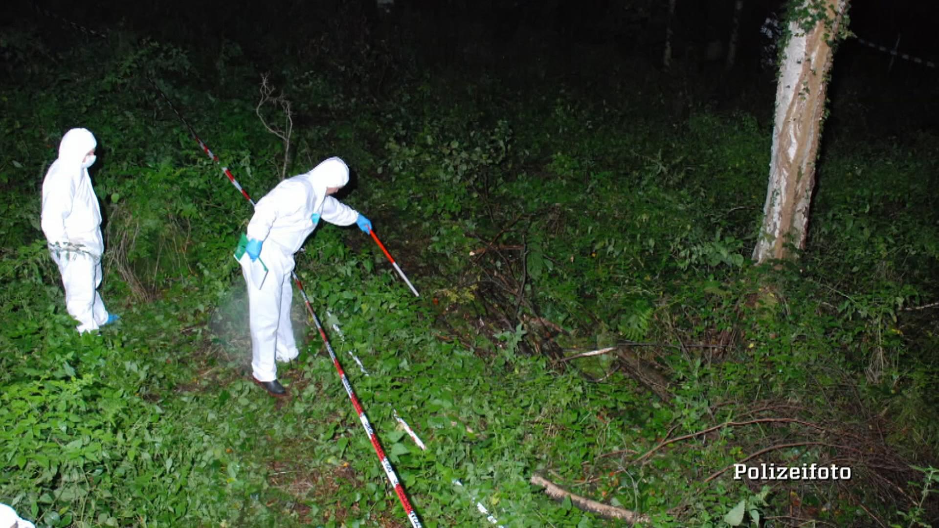 Zwei Ermittler in weißen Kriminaltechnikeranzügen durchsuchen nachts einen Haufen Äste in einem Wald. Der eine Ermittler tastet sich mit einem weiß-roten-Stock hinter einer Flatterbandabsperrung vor. Der andere Ermittler beobachtet von hinten die Szenerie. Die Markierung "Polizeifoto" liegt darüber.