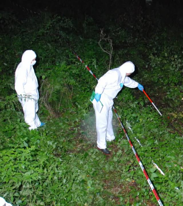 Zwei Ermittler in weißen Kriminaltechnikeranzügen durchsuchen nachts einen Haufen Äste in einem Wald. Der eine Ermittler tastet sich mit einem weiß-roten-Stock hinter einer Flatterbandabsperrung vor. Der andere Ermittler beobachtet von hinten die Szenerie. Die Markierung "Polizeifoto" liegt darüber.