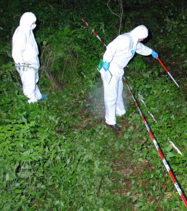 Zwei Ermittler in weißen Kriminaltechnikeranzügen durchsuchen nachts einen Haufen Äste in einem Wald. Der eine Ermittler tastet sich mit einem weiß-roten-Stock hinter einer Flatterbandabsperrung vor. Der andere Ermittler beobachtet von hinten die Szenerie. Die Markierung "Polizeifoto" liegt darüber.