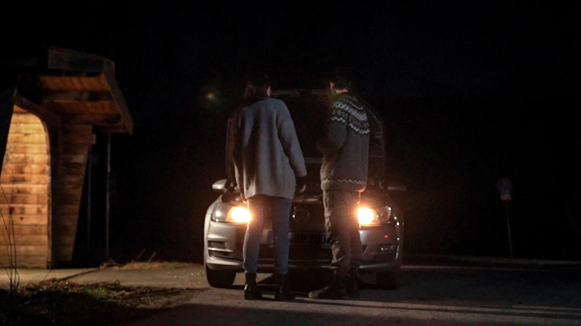 Es ist Nacht. An der Seite steht ein Wartehäuschen aus Holz einer Bushaltestelle. In dem Häuschen brennt Licht. Neben dem Haltestellenhaus steht ein dunkelgrauer Pkw dessen Scheinwerfer leuchten. Die Motorhaube des Wagens ist geöffnet. Vor dem Wagen stehen zwei Personen, links eine Frau, rechts daneben ein Mann. Sie blicken aud die offen stehende Motorhaube.