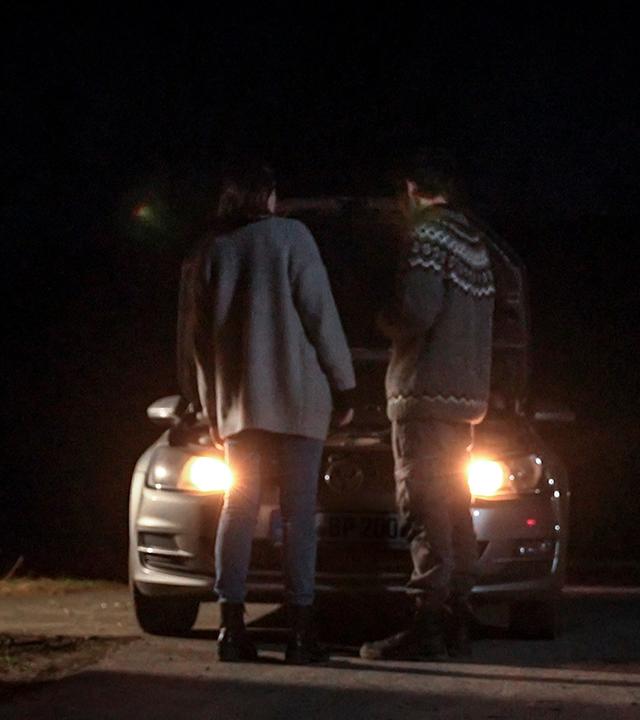 Es ist Nacht. An der Seite steht ein Wartehäuschen aus Holz einer Bushaltestelle. In dem Häuschen brennt Licht. Neben dem Haltestellenhaus steht ein dunkelgrauer Pkw dessen Scheinwerfer leuchten. Die Motorhaube des Wagens ist geöffnet. Vor dem Wagen stehen zwei Personen, links eine Frau, rechts daneben ein Mann. Sie blicken aud die offen stehende Motorhaube.