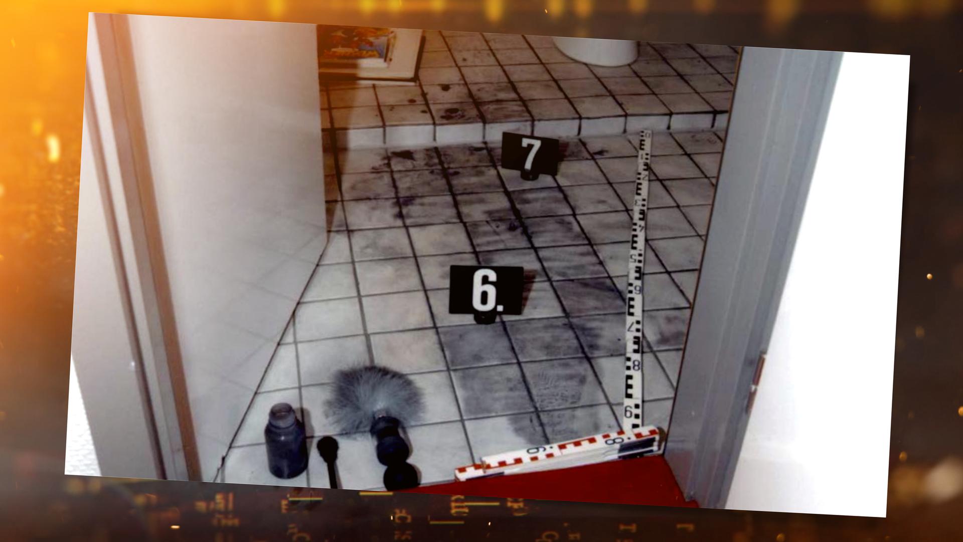 Blick von oben durch die Tür in ein Badezimmer: Auf dem gefliesten Boden sind dunkle Spuren von Fingerabdruckpulver verteilt. Zwei schwarze Spurenschilder mit den Nummern "6" und "7" markieren Fundstellen. Am Rand liegt ein Maßstab der Spurensicherung im Türrahmen. Im Vordergrund liegen Arbeitsutensilien der Spurensicherung: ein Pinsel mit grauen Borsten, ein Blasebalg sowie ein Behälter mit Pulver. Im Hintergrund liegt ein aufgeschlagenes Buch am Boden neben einer Toilette.