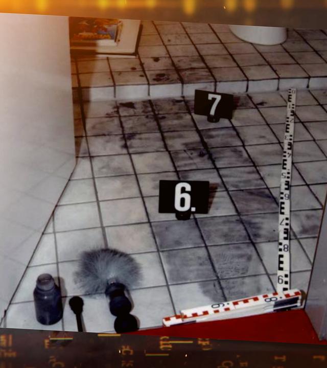 Blick von oben durch die Tür in ein Badezimmer: Auf dem gefliesten Boden sind dunkle Spuren von Fingerabdruckpulver verteilt. Zwei schwarze Spurenschilder mit den Nummern "6" und "7" markieren Fundstellen. Am Rand liegt ein Maßstab der Spurensicherung im Türrahmen. Im Vordergrund liegen Arbeitsutensilien der Spurensicherung: ein Pinsel mit grauen Borsten, ein Blasebalg sowie ein Behälter mit Pulver. Im Hintergrund liegt ein aufgeschlagenes Buch am Boden neben einer Toilette.
