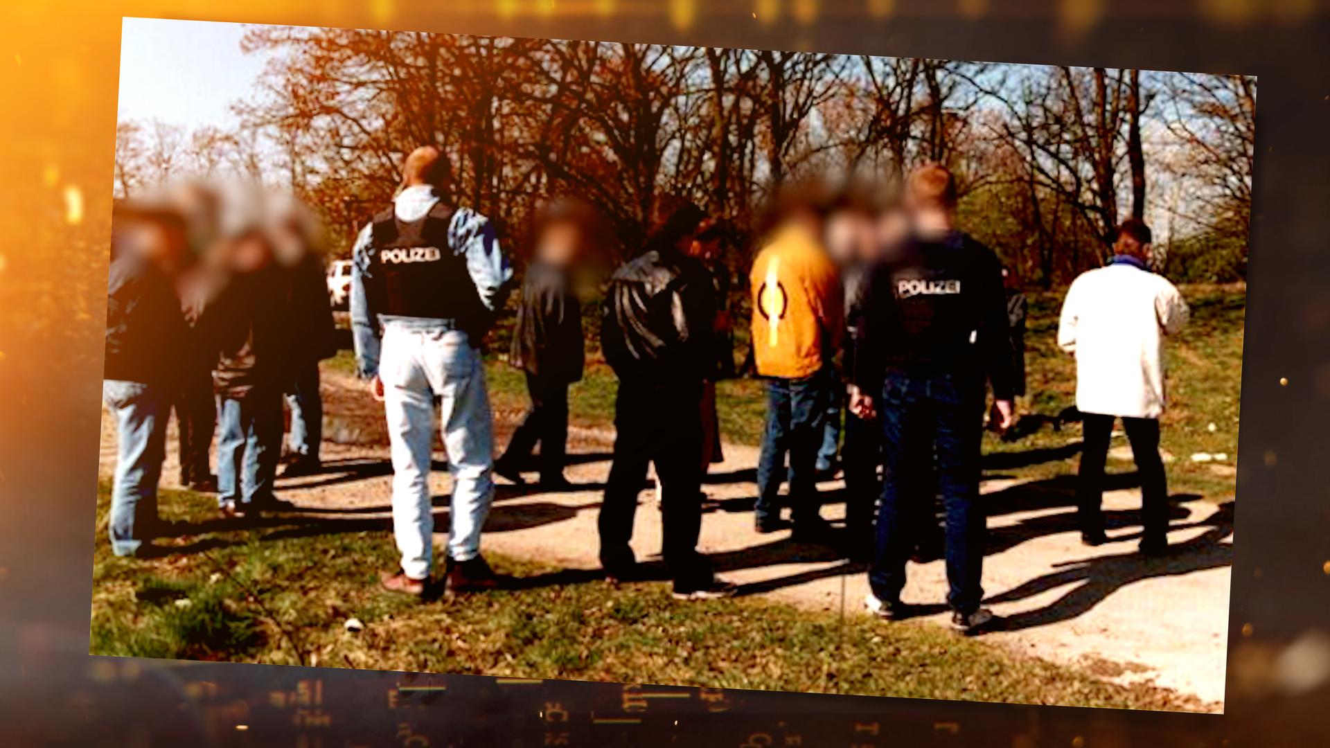 Mehrere Personen stehen auf einem schmalen Feldweg zwischen Wiese und Bäumen. Einige tragen Westen mit der Aufschrift "Polizei". Alle Personen stehen mit dem Rücken zur Kamera. Die Gesichter sind unkenntlich gemacht