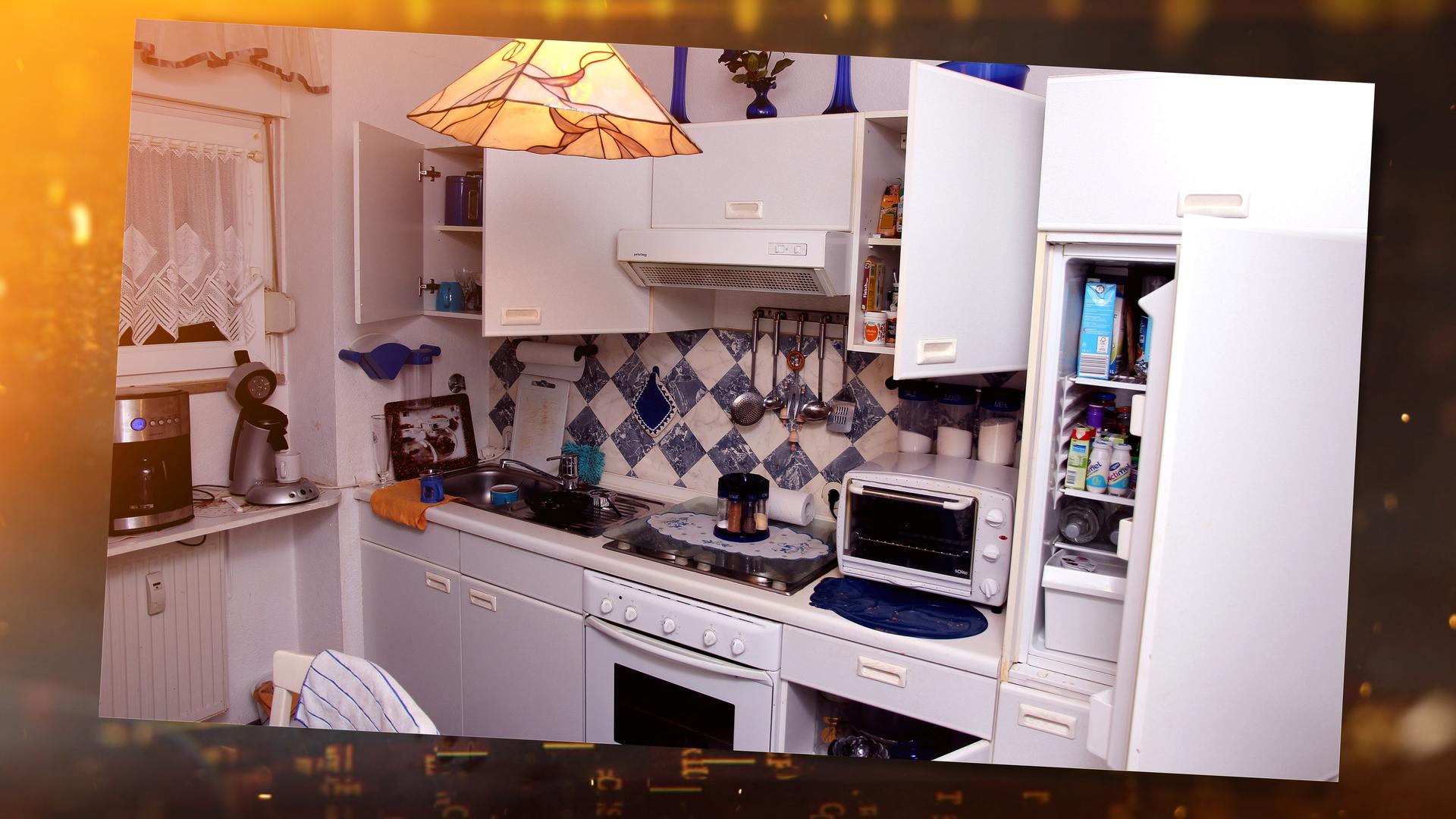 In einer weißen Einbauküche, die sehr aufgeräumt wirkt, sind Ober- und Unterschränke teilweise geöffnet. Im geöffneten Kühlschrank an der Seite sieht man verschiedene Getränke und Joghurtpackungen.