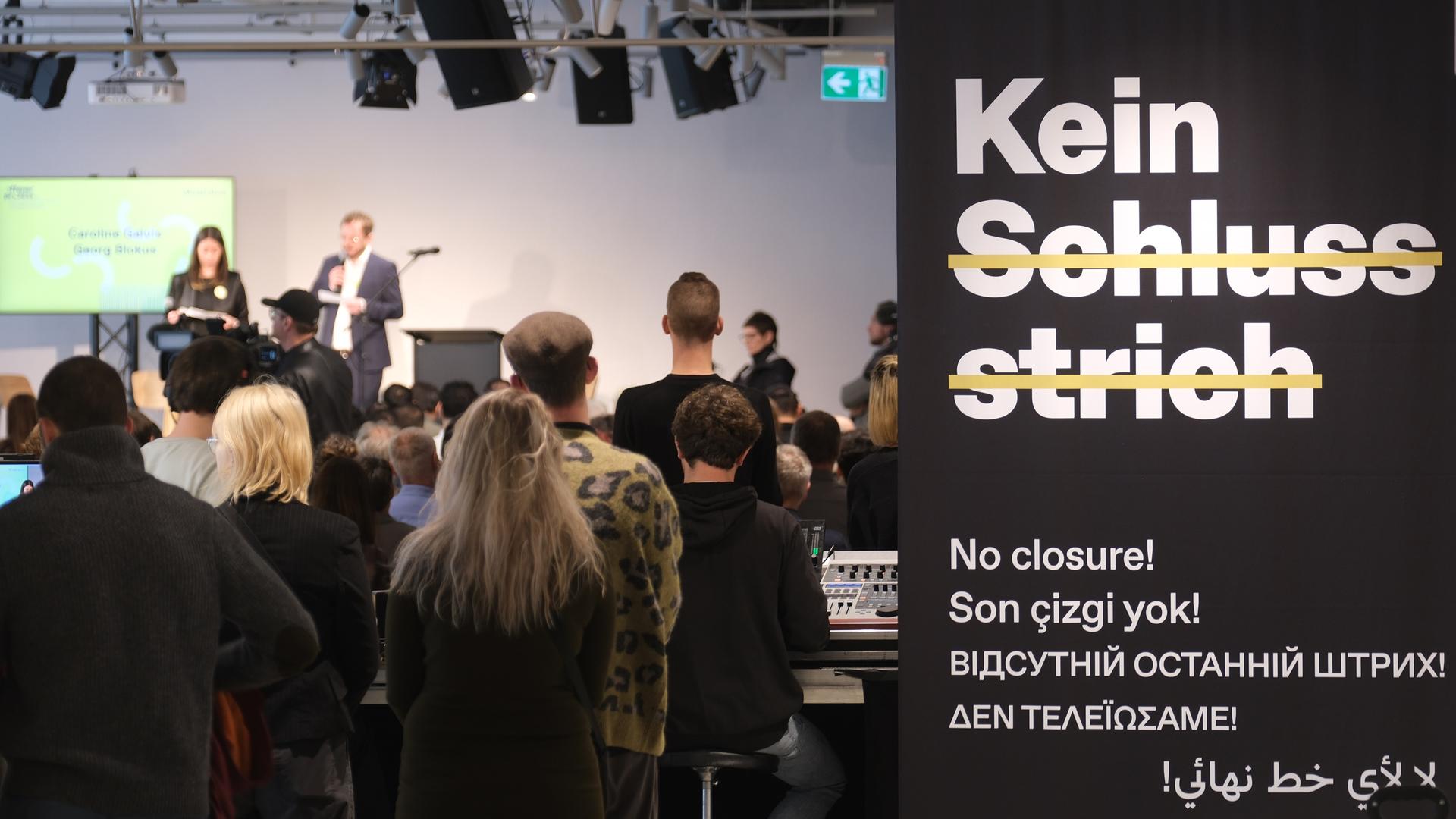 Die Eröffung des Dokumentationszentrums. Zwei Menschen sprechen auf einer Bühne, viele andere Menschen stehen davor. Im rechten Bildrand sind Ausschnitte der Ausstellung zu sehen.