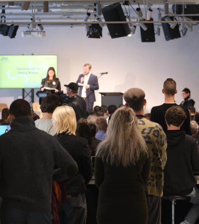 Die Eröffung des Dokumentationszentrums. Zwei Menschen sprechen auf einer Bühne, viele andere Menschen stehen davor. Im rechten Bildrand sind Ausschnitte der Ausstellung zu sehen.