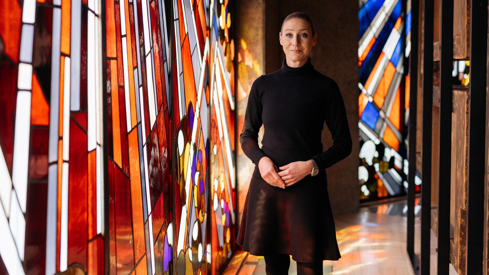 Alexandra Winkel steht in einer Kirche, sie lächelt sanft in die Kamera. Im Hintergrund sind bunte Kirchenfenster zu sehen.