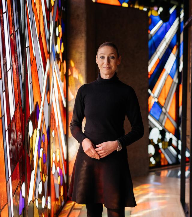 Alexandra Winkel steht in einer Kirche, sie lächelt sanft in die Kamera. Im Hintergrund sind bunte Kirchenfenster zu sehen.