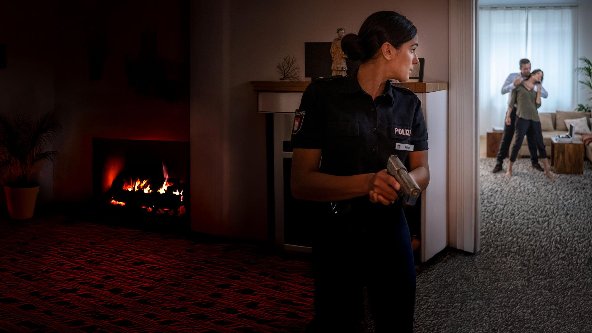 "Notruf Hafenkante - Erster Einsatz": Pinar Aslan (Aybi Era) steht versteckt neben einer geöffneten Wohnzimmertür und blickt vorsichtig ins Wohnzimmer. Dort steht Dr. Jasmin Jonas (Gerit Kling). Hinter ihr steht ein Mann. Er hat seinen linken Arm um ihren Hals gelegt. In der rechten Hand hält er eine Spritze.
