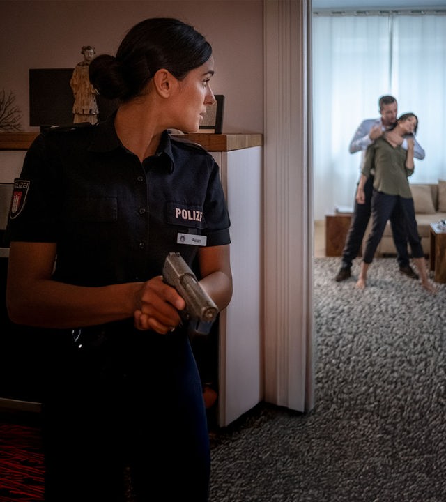 "Notruf Hafenkante - Erster Einsatz": Pinar Aslan (Aybi Era) steht versteckt neben einer geöffneten Wohnzimmertür und blickt vorsichtig ins Wohnzimmer. Dort steht Dr. Jasmin Jonas (Gerit Kling). Hinter ihr steht ein Mann. Er hat seinen linken Arm um ihren Hals gelegt. In der rechten Hand hält er eine Spritze.