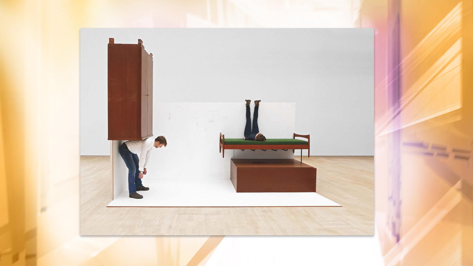 Das in einem großen Museumsraum ausgestellte Kunstwerk besteht aus einem Schrank, der verkehrt herum an der Wand hängt. Unter ihm beugt sich ein Mann vor, als ob er den Schrank auf dem Rücken tragen würde. An der Wand daneben hängt ein Bett, auf dem eine Person quer liegt und die Beine an der Wand hoch streckt. Darunter liebt ein Schrank quer.